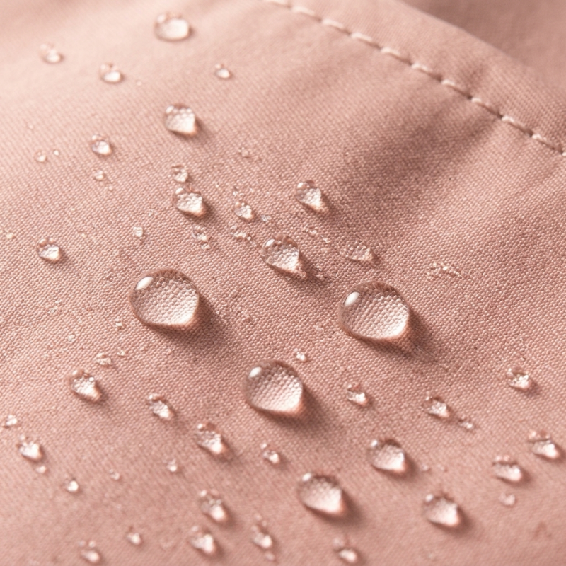 Close-up of a dusty pink fabric surface with water droplets beading on top, showing a smooth, slightly textured material with fine stitching.