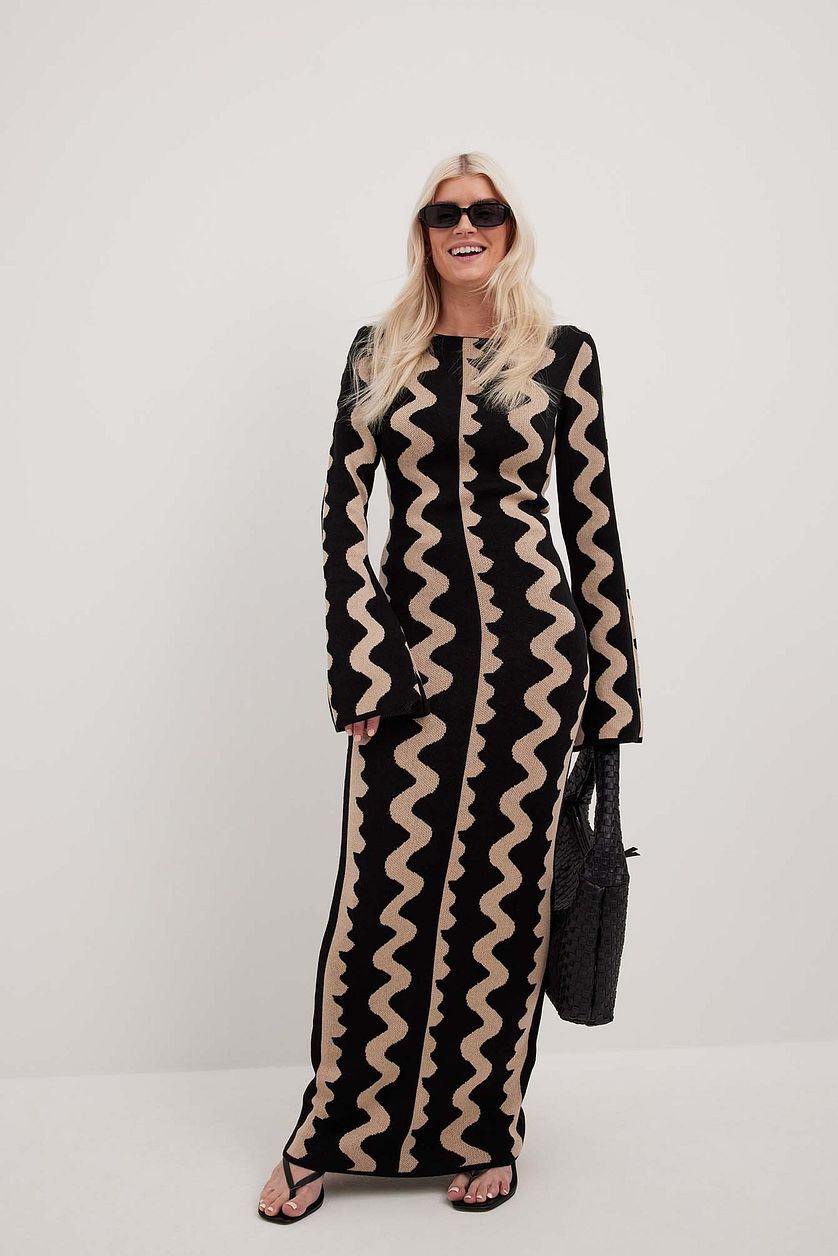 Model laughs head to toe photography of Gemini Knit Dress by ShoptheKei with a black leather tote bag and oval shaped black sunglasses. Sweater dress maxi length with long flare sleeves. Body conscious. Mod 1960's style dress with vertical wavy squiggle line patterns.