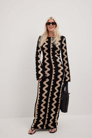 Model laughs head to toe photography of Gemini Knit Dress by ShoptheKei with a black leather tote bag and oval shaped black sunglasses. Sweater dress maxi length with long flare sleeves. Body conscious. Mod 1960's style dress with vertical wavy squiggle line patterns.