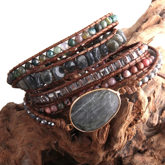Gray Bohemian style wrap bracelet with a large center stone and several colorful smaller stones set in a natural cording. The bracelet rests on driftwood. Gray Boho Crystal Bracelet at Shop the Kei