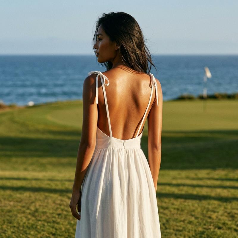 The open back view of a woman in a white dress standing on a golf course with ocean in the background