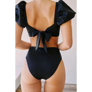 Back view of model standing in the black Lylah Bikini from ShoptheKei.com. Short sleeves are puffed and top straps are tied into a bow, to show the low back neckline. The high waist bikini bottom and high cut legs are visible.