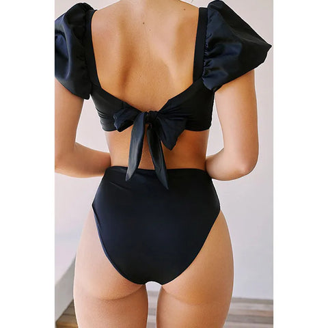 Back view of model standing in the black Lylah Bikini from ShoptheKei.com. Short sleeves are puffed and top straps are tied into a bow, to show the low back neckline. The high waist bikini bottom and high cut legs are visible.