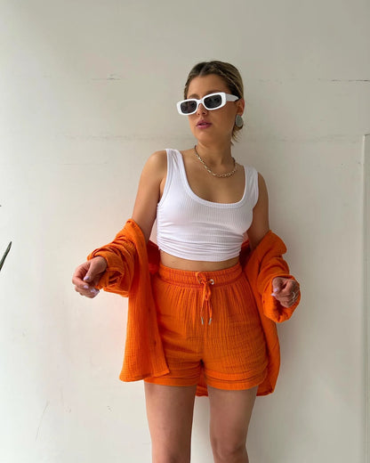 Colada Shorts Set from Shop the Kei - Model wearing a vibrant orange matching shorts set featuring a textured fabric with subtle vertical ribbing. The set includes loose-fitting shorts with drawstring waist and an unbuttoned long-sleeve shirt worn open over the forearms with a white ribbed crop top. The outfit is accessorized with white framed sunglasses, a simple gold chain necklace and rings. Photographed against a plain white background