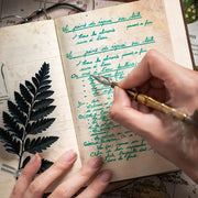 Hand writing in a notebook with a quill pen, surrounded by nature-themed elements.