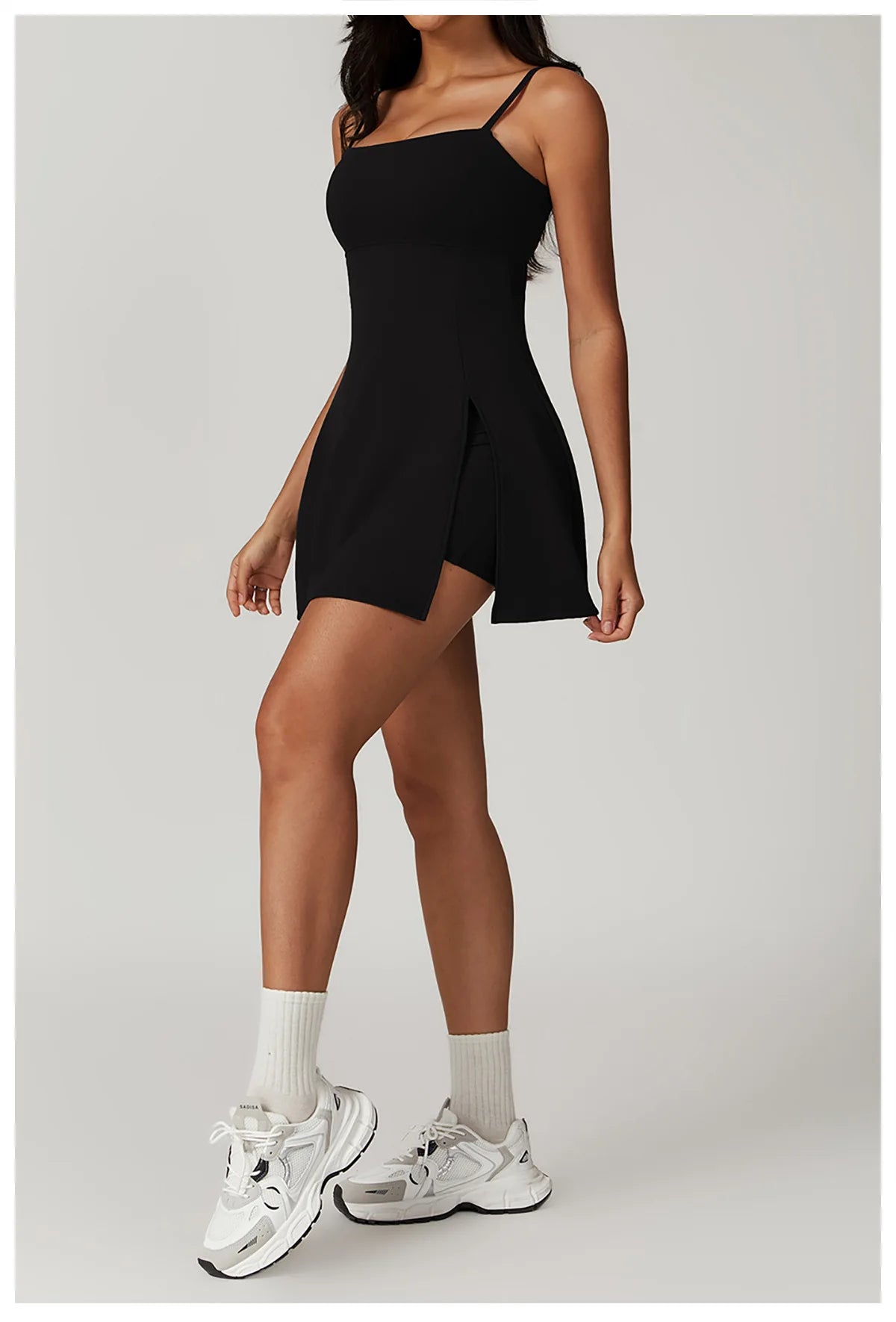 Side view of the Bree Yoga Dress in classic black color. One uplifted knee reveals a slit in the skirt and hidden shorts. Model styled with white ankle socks and chunky athletic sneakers against a neutral background.