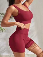 Model wearing Zen Yoga Shorts Set in red dust. Soft textured rib knit tank top with matching slim fit high waist shorts provide support and comfort during your movements. Model stands to the side with a hand on her waist and the other rests at her head in front of a white background.