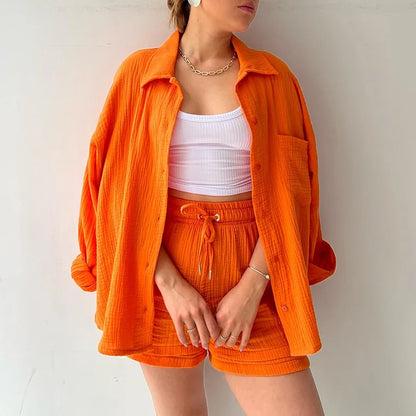 Colada Shorts Set from Shop the Kei - Model wearing a vibrant orange matching shorts set featuring a textured fabric with subtle vertical ribbing. The set includes loose-fitting shorts with drawstring waist and an unbuttoned long-sleeve shirt worn open over a white ribbed crop top. The outfit is accessorized with a simple gold chain necklace and rings. Photographed against a plain white background