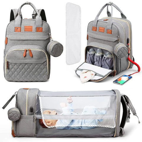 Gray Lotus 3-in-1 Bassinet Diaper Bag backpack with orange leather accents, featuring quilted front pocket, dual carry handles, and small circular accessory pouch. Front of bag is displayed closed. A quarter turn of the bag reveals a thermal bottle pocket unzipped. Fully extended bassinet feature displayed with a baby inside