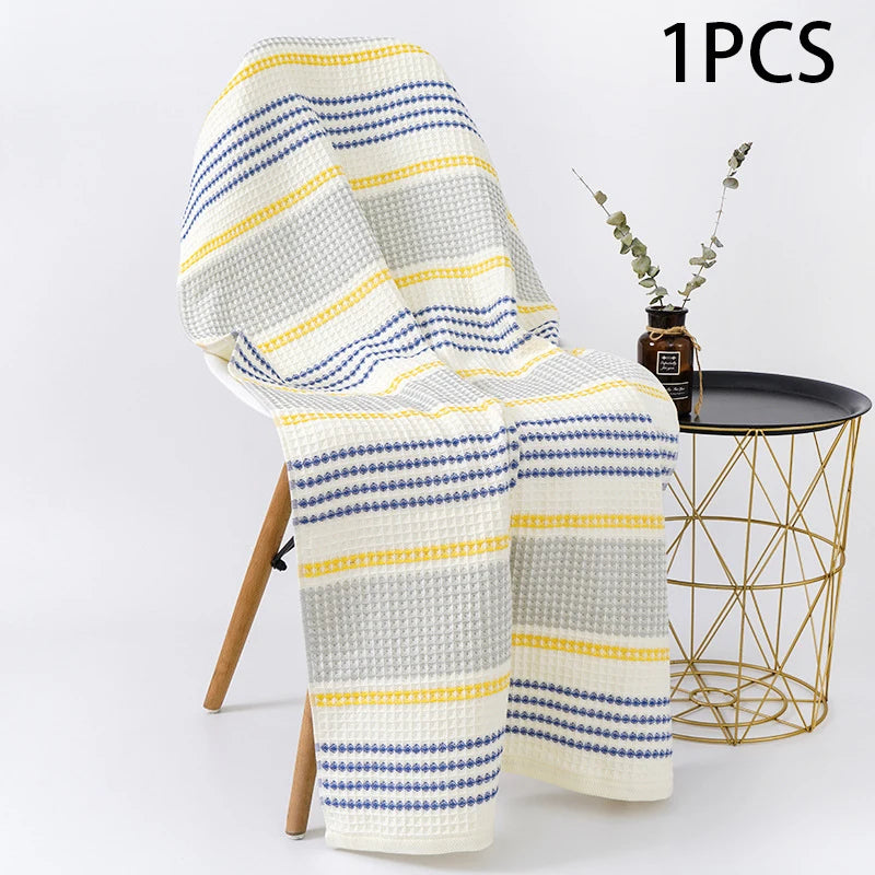 Lemonade PetalDry Striped Cotton Bath Towel, in yellow, gray, and navy stripes, draped over a wooden chair beside a gold wire side table with a eucalyptus stem.