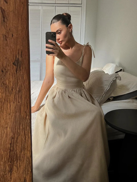 Isla Summer Dress from Shop the Kei - Brunette model wears a natural linen dress with gathered waist, v-neckline, and tied spaghetti straps. She sits on a bed photographed with a phone in her hand