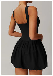 Back view of a model wearing the black Rinay Sport Dress from ShoptheKei activewear collection. Sleeveless tank dress with a low square shaped back neckline, gathered skirt at waist seam, and blouson hem. Stretchy and cute dress design for sports and casual strolls.