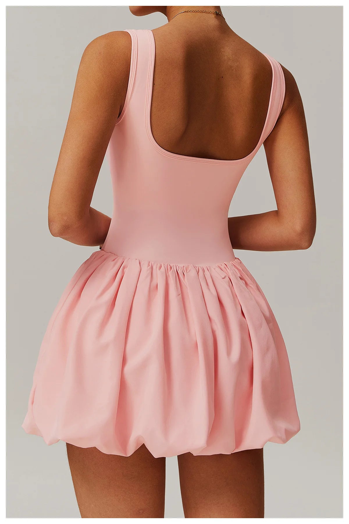 Back view of a model wearing the pink Rinay Sport Dress from ShoptheKei activewear collection. Sleeveless tank dress with a low square shaped back neckline, gathered skirt at waist seam, and blouson hem. Stretchy and cute dress design for sports and casual strolls.