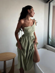 Isla Summer Dress from Shop the Kei - Sage green linen dress with gathered waist, worn by a woman standing near a window with a wooden side table, showcasing elegant and minimalist design
