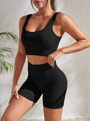 Model wearing Zen Yoga Shorts Set in black. Soft textured rib knit tank top with matching slim fit high waist shorts provide support and comfort during your movements. Model stands ¾ with a hand at her waist and the other rests on her leg in front of a white background.