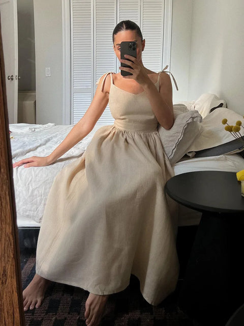 Isla Summer Dress from Shop the Kei - Brunette model wears a natural linen dress with gathered waist, v-neckline, and tied spaghetti straps. She sits on a bed photographed with a phone in her hand