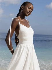 Isla Summer Dress from Shop the Kei - Soft white linen dress with gathered waist, worn by a woman standing against the cloudy sky and shore. Paired with minimalist jewelry and long beaded braids