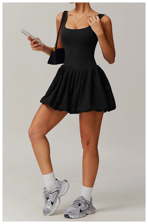 Full length front view of a model wearing the black Rinay Sport Dress from ShoptheKei activewear collection. Sleeveless tank dress with gathered skirt at waist seam and blouson hem. Stretchy and cute dress design for sports and casual strolls. Paired with crew socks and gray sneakers.