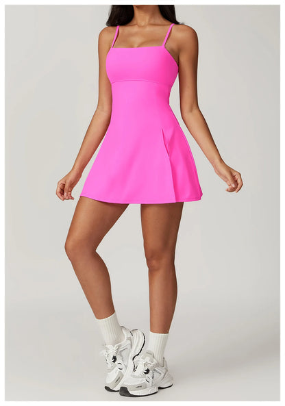 Front view of the Bree Yoga Dress in "Bubblegum pink" color. A mini dress with thin spaghetti straps, straight neckline, fitted bodice, and short A-line skirt. Model poses with white ankle socks and chunky athletic sneakers against a neutral background.
