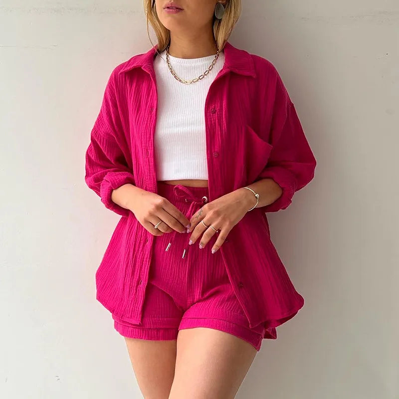 Colada Shorts Set from Shop the Kei - Model wearing a vibrant hot pink matching shorts set featuring a textured fabric with subtle vertical ribbing. The set includes loose-fitting shorts with drawstring waist and an unbuttoned long-sleeve shirt worn open over a white ribbed crop top. The outfit is accessorized with a simple gold chain necklace and rings. Photographed against a plain white background