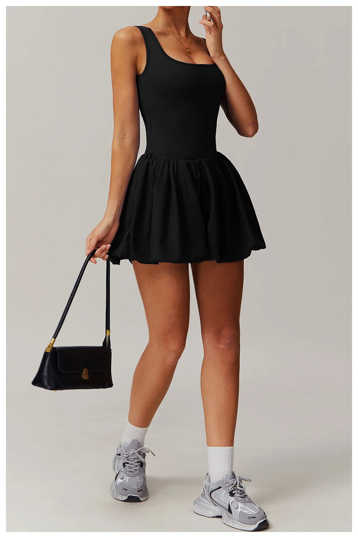 Full length front view of a model wearing the black Rinay Sport Dress from ShoptheKei activewear collection. Sleeveless tank dress with gathered skirt at waist seam and blouson hem. Stretchy and cute dress design for sports and casual strolls. Paired with a black purse, crew socks, and gray sneakers.