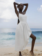 Isla Summer Dress from Shop the Kei - Soft white linen dress with gathered waist, worn by a woman standing on the sand against the cloudy sky and shore. Paired with minimalist jewelry and long beaded braids