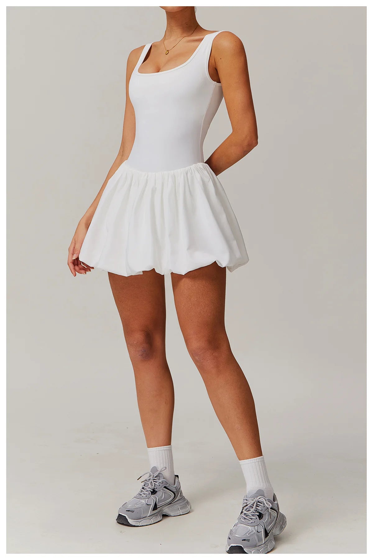 Full length front view of a model wearing the white Rinay Sport Dress from ShoptheKei activewear collection. Sleeveless tank dress with gathered skirt at waist seam and blouson hem. Stretchy and cute dress design for sports and casual strolls. Paired with crew socks and gray sneakers.