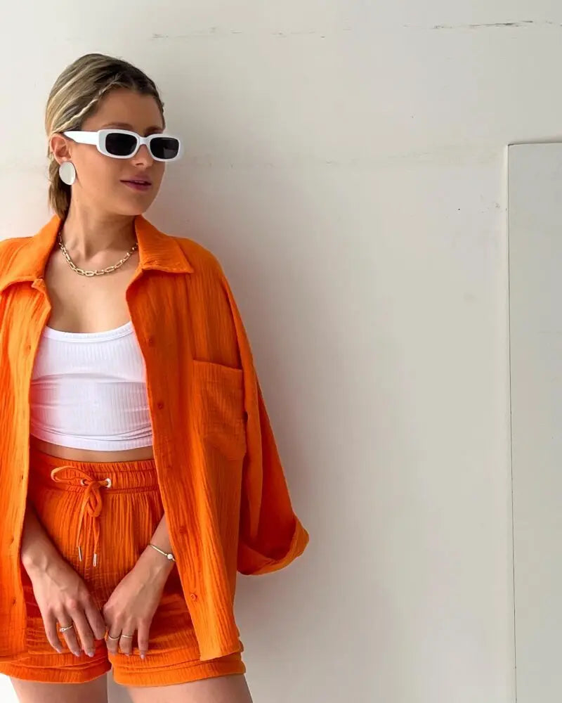 Colada Shorts Set from Shop the Kei - Model wearing a vibrant orange matching shorts set featuring a textured fabric with subtle vertical ribbing. The set includes loose-fitting shorts with drawstring waist and an unbuttoned long-sleeve shirt worn open over a white ribbed crop top. The outfit is accessorized with white framed sunglasses, a simple gold chain necklace and rings. Photographed against a plain white background