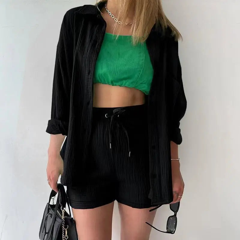 Colada Shorts Set from Shop the Kei - Model wearing a classic black matching shorts set featuring a textured fabric with subtle vertical ribbing. The set includes loose-fitting shorts with drawstring waist and an unbuttoned long-sleeve shirt worn open over a green linen crop top. The outfit is accessorized with a simple gold chain necklace, black sunglasses and purse. Photographed against a plain white background