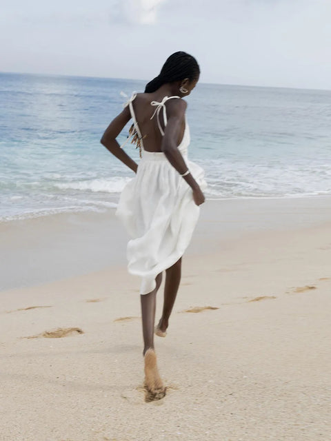 Isla Summer Dress from Shop the Kei - Soft white linen dress with gathered waist and tied spaghetti straps, worn by a woman leaping on the beach against a blue sky and shore. Paired with minimalist jewelry, long beaded braids, and joy!