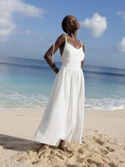 Isla Summer Dress from Shop the Kei - Soft white linen dress with gathered waist, worn by a woman standing on the sand against the cloudy sky and shore. Paired with silver minimalist jewelry