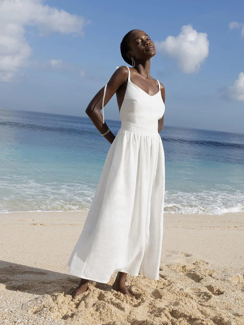 Isla Summer Dress from Shop the Kei - Soft white linen dress with gathered waist, worn by a woman standing on the sand against the cloudy sky and shore. Paired with silver minimalist jewelry