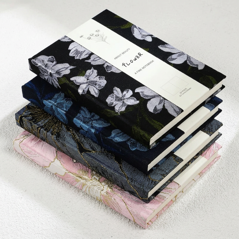 A stack of four decorative notebooks with floral and marbled covers, arranged diagonally on a textured white surface. The top notebook is black with white flowers and a white paper band labeled 'Flower' and 'A Fine Notebook.