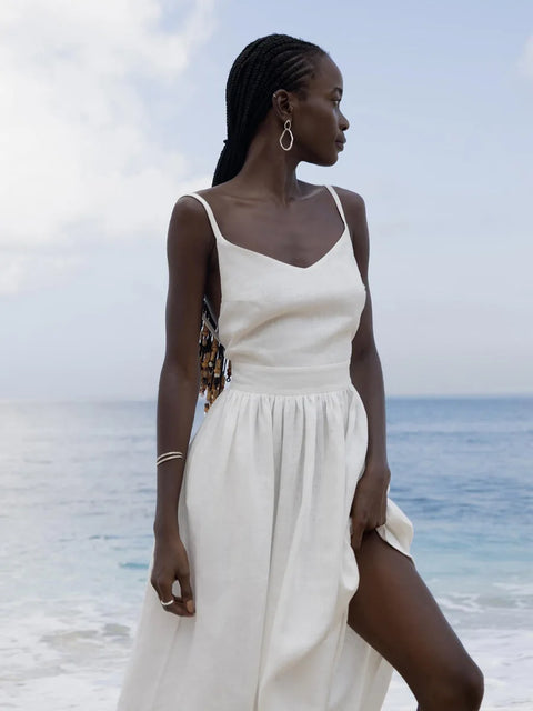 Isla Summer Dress from Shop the Kei - Soft white linen dress with gathered waist, worn by a woman standing against the cloudy sky and crystal blue shore. Paired with minimalist jewelry and long beaded braids