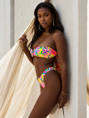 Side view of model poses in a floating cabana wearing Miami tropical Cali Bandeau Bikini Set from ShoptheKei.com swim collection. Electric yellow, pink, orange, and green floral bandeau bikini with an O-ring, low-waist bottoms, and wire-free padded support for a chic beach look.