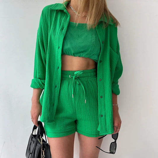 Colada Shorts Set from Shop the Kei - Model wearing a vibrant green matching shorts set featuring a textured fabric with subtle vertical ribbing. The set includes loose-fitting shorts with drawstring waist and an unbuttoned long-sleeve shirt worn open over a green linen crop top. The outfit is accessorized with a simple gold chain necklace, black sunglasses and purse. Photographed against a plain white background