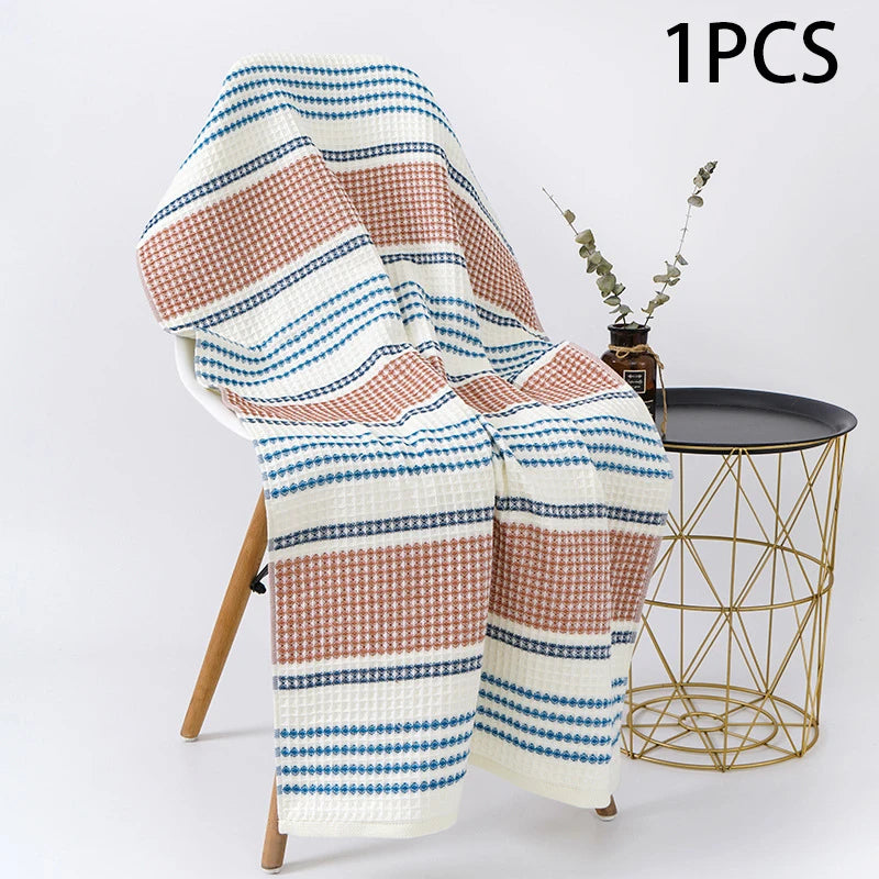 Earthy PetalDry Striped Cotton Bath Towel, in terracotta and navy stripes, draped over a wooden chair beside a gold wire side table with a eucalyptus stem.