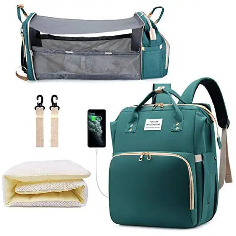 Green baby diaper backpack shown in multiple views: with additional straps, with USB charging port, folded mattress insert, and fully expanded as bassinet with mesh sides containing a baby. Lotus 3-in-1 bassinet diaper bag travel system with partial canopy