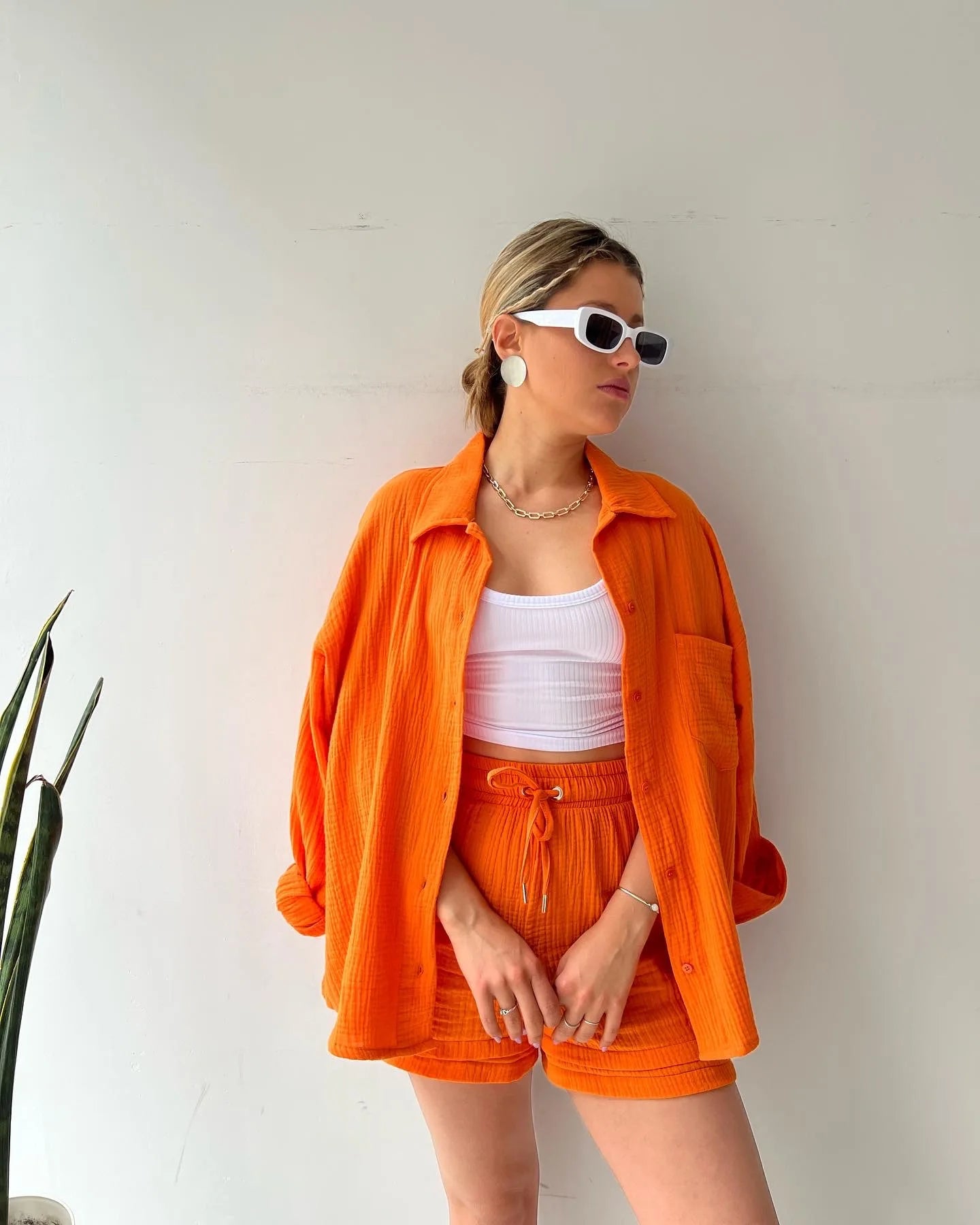 Colada Shorts Set from Shop the Kei - Model wearing a vibrant orange matching shorts set featuring a textured fabric with subtle vertical ribbing. The set includes loose-fitting shorts with drawstring waist and an unbuttoned long-sleeve shirt worn open over a white ribbed crop top. The outfit is accessorized with white framed sunglasses, a simple gold chain necklace and rings. Photographed against a plain white background