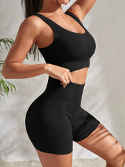 Model wearing Zen Yoga Shorts Set in black. Soft textured rib knit tank top with matching slim fit high waist shorts provide support and comfort during your movements. Model stands to the side with a hand on her waist and the other rests at her head in front of a white background.