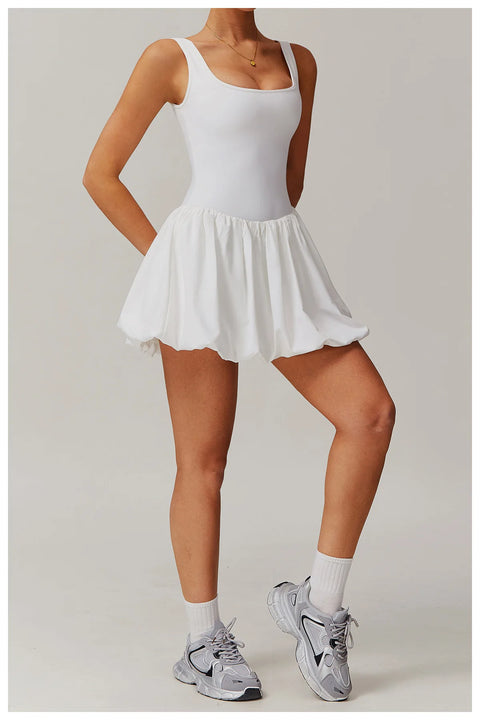 Full length front view of a model wearing the white Rinay Sport Dress from ShoptheKei activewear collection. Sleeveless tank dress with gathered skirt at waist seam and blouson hem. Stretchy and cute dress design for sports and casual strolls. Paired with crew socks and gray sneakers.