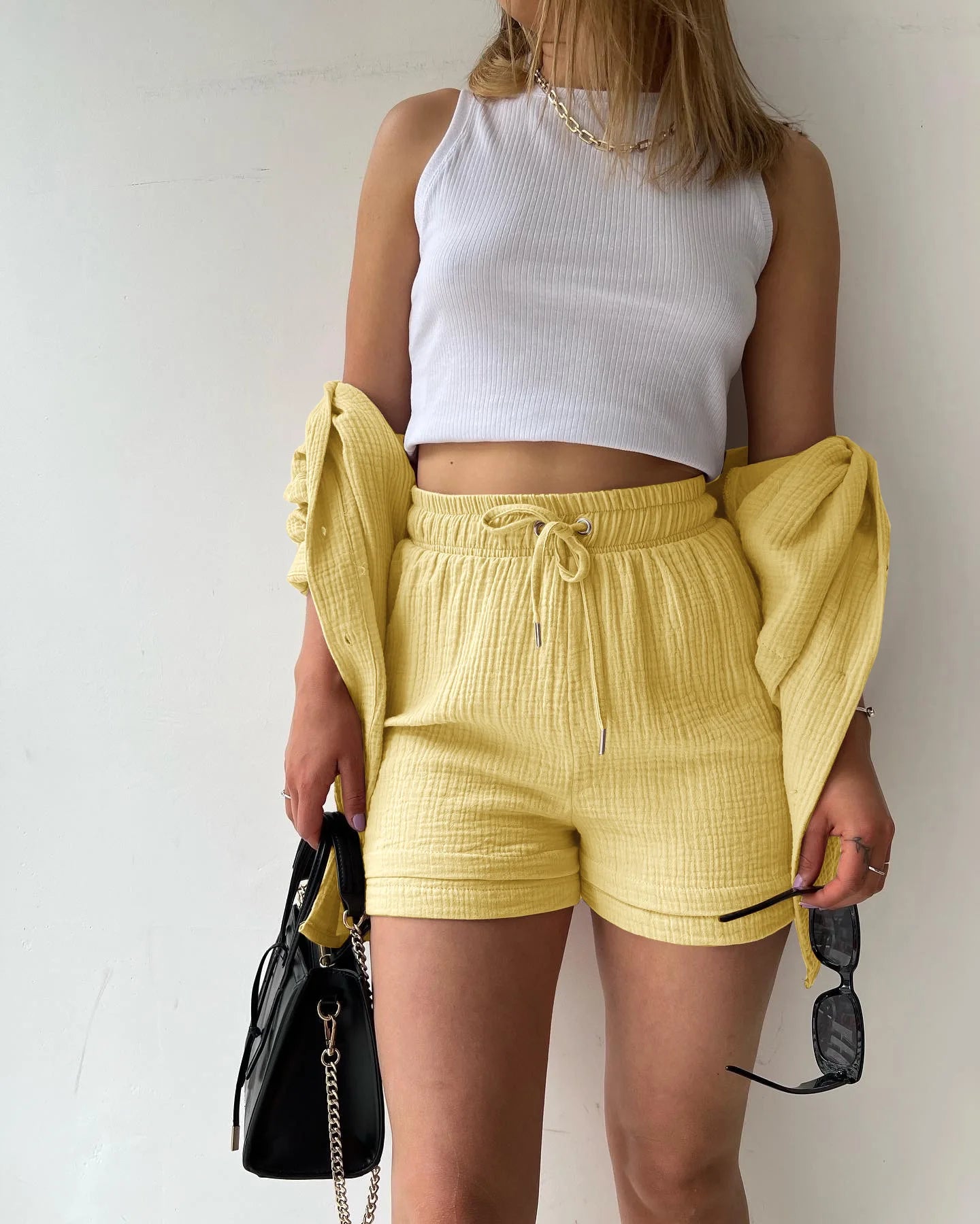 Colada Shorts Set from Shop the Kei - Model wearing a buttery yellow matching shorts set featuring a textured fabric with subtle vertical ribbing. The set includes loose-fitting shorts with drawstring waist and an unbuttoned long-sleeve shirt worn open over a white ribbed crop top. The outfit is accessorized with a simple gold chain necklace and rings. Photographed against a plain white background