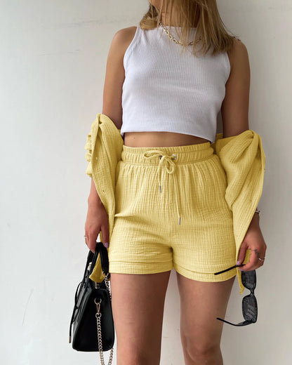 Colada Shorts Set from Shop the Kei - Model wearing a buttery yellow matching shorts set featuring a textured fabric with subtle vertical ribbing. The set includes loose-fitting shorts with drawstring waist and an unbuttoned long-sleeve shirt worn open over a white ribbed crop top. The outfit is accessorized with a simple gold chain necklace and rings. Photographed against a plain white background