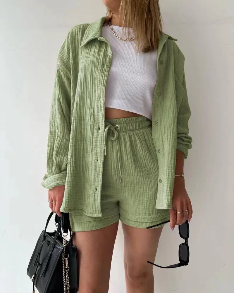 Colada Shorts Set from Shop the Kei - Model wearing a soft green matching shorts set featuring a textured fabric with subtle vertical ribbing. The set includes loose-fitting shorts with drawstring waist and an unbuttoned long-sleeve shirt worn open over a white ribbed crop top. The outfit is accessorized with a simple gold chain necklace, black sunglasses, and purse. Photographed against a plain white background