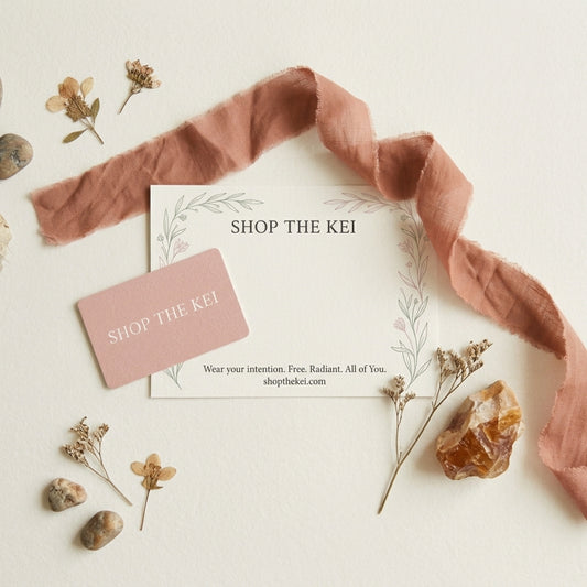 A styled flat-lay featuring a white card and a pink contact card both printed with the brand name 'SHOP THE KEI', surrounded by a dusty rose ribbon, dried flowers, small stones, and a polished amber crystal on a warm off-white background.