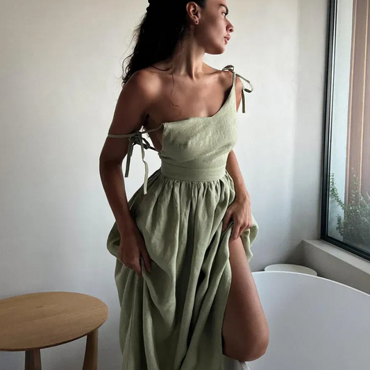 Isla Summer Dress from Shop the Kei - Sage green linen dress with gathered waist, worn by a woman standing near a window with a wooden side table, showcasing elegant and minimalist design