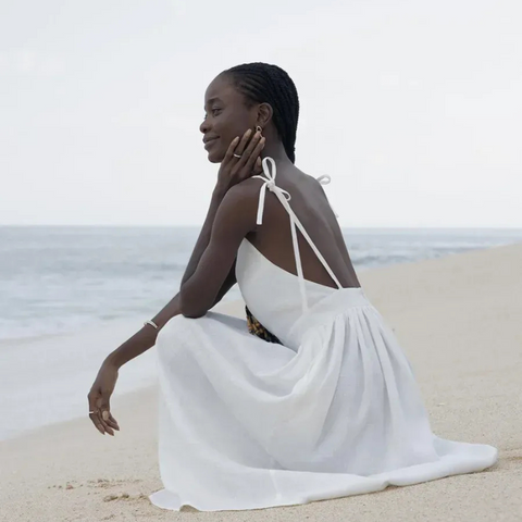 Isla Summer Dress from Shop the Kei - Soft white linen dress with gathered waist, worn by a woman sitting on the sand gazing peacefully at the shore. Paired with minimalist jewelry and long beaded braids
