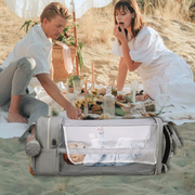A couple having a beach picnic with a Lotus 3-in-1 Bassinet Diaper Bag in the foreground. The couple is dressed in white, sitting on a white blanket with a picnic spread between them including food and drinks on a sandy beach. A baby is visible inside the bassinet portion of the diaper bag which is gray with mesh sides. The setting appears to be a sunny day with golden grasses in the background
