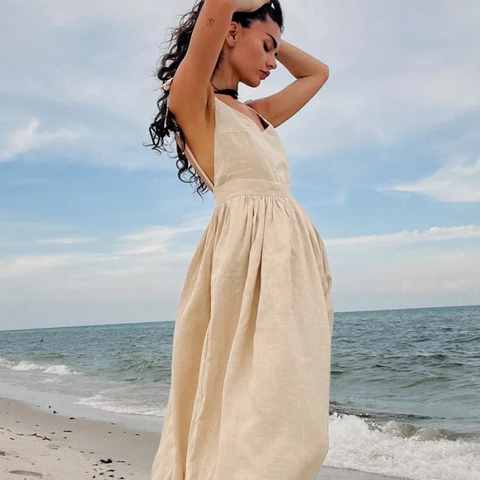 Isla Summer Dress from Shop the Kei - Natural linen dress with gathered waist, worn by a woman standing on the sand gazing peacefully at the shore.