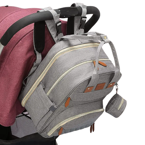 Gray Lotus 3-in-1 Bassinet Diaper Bag backpack with orange leather accents, featuring quilted front pocket, dual carry handles, and small circular accessory pouch. Bag is hanging on the handles of a stroller